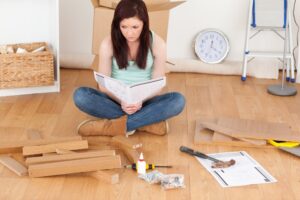Woman reading furniture assembly instructions while sitting cross-legged on floor surrounded by wooden parts and tools